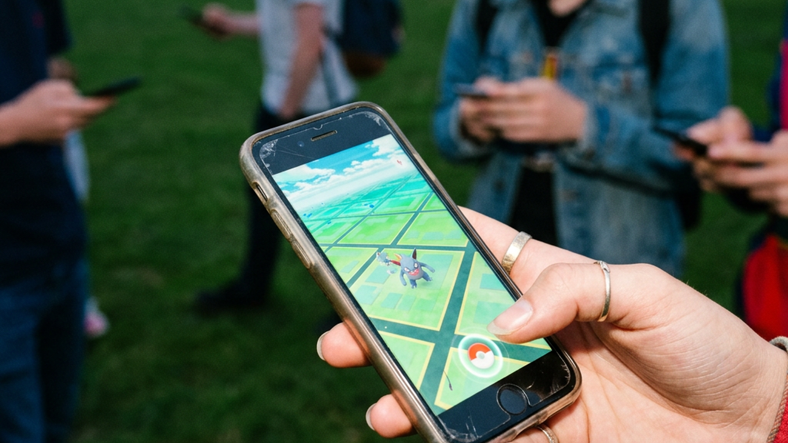 A hand holding a cracked phone playing an augmented-reality monster-catching game while a group plays in the same park